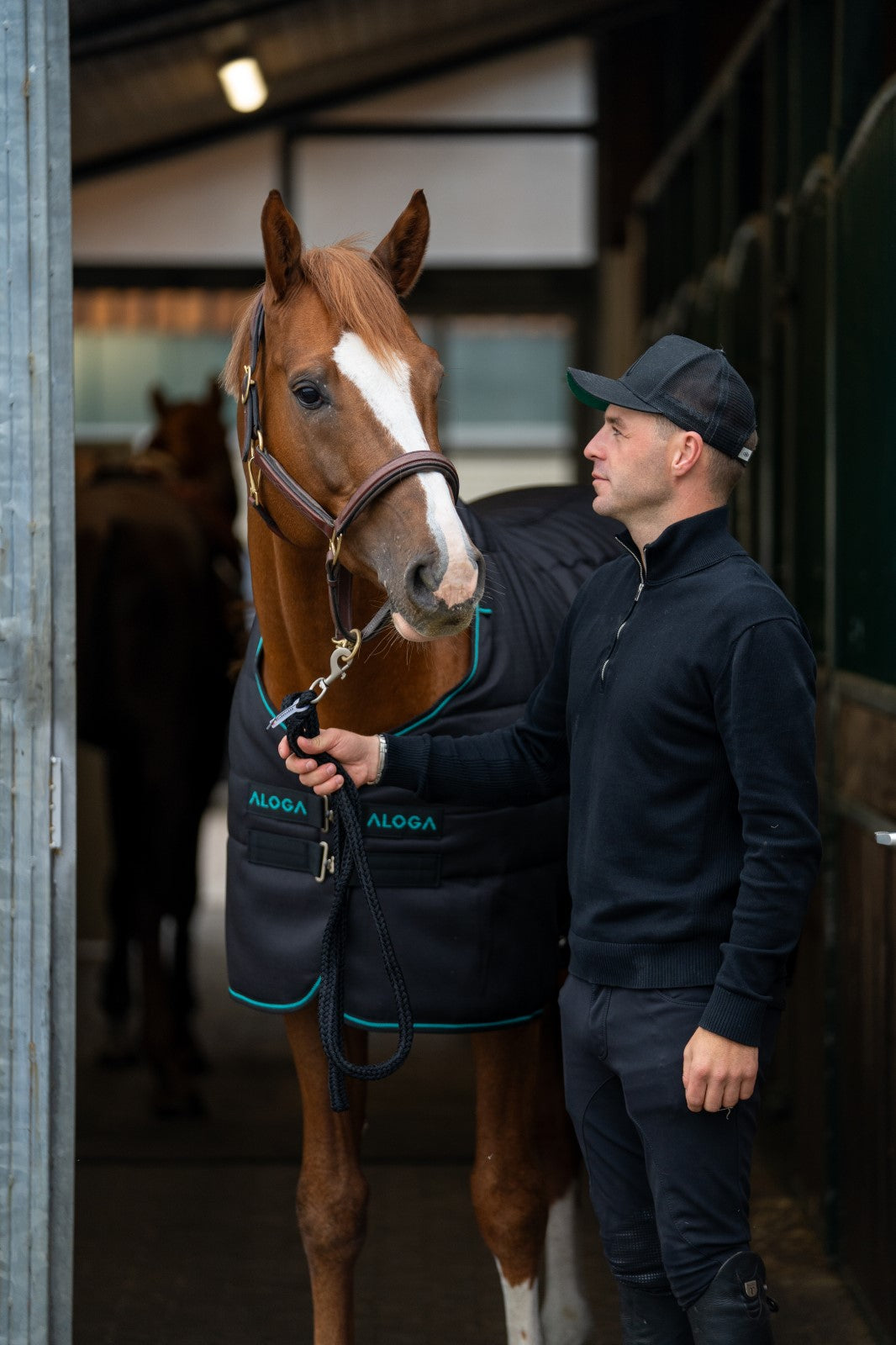 Man standing next to a horse wearing a black ALGORA blanket in a stable.